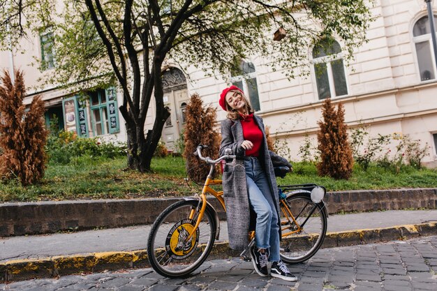 Het portret van gemiddelde lengte van modieuze vrouwelijke student in uitstekende jeans die met gele fiets stellen