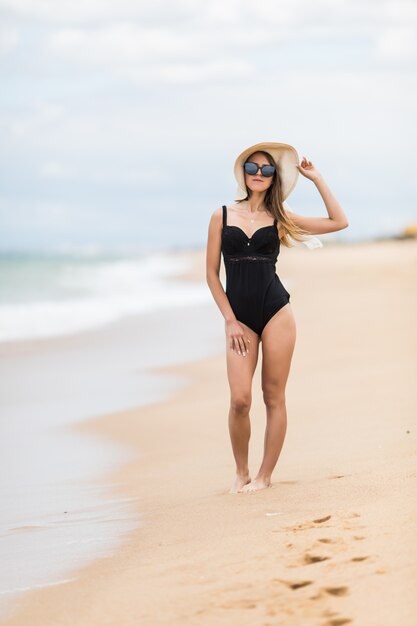 Het portret van gemiddelde lengte van schitterende jonge vrouw die in strohoed op zandig strand loopt