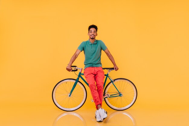 Het portret van gemiddelde lengte van zekere Afrikaanse mens die zich voor zijn fiets bevindt. emotionele zwarte man in lichte outfit poseren met fiets.