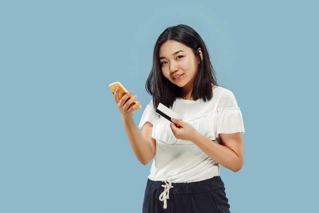 Het portret van halve lengte van de Koreaanse jonge vrouw. Vrouwelijk model dat haar smartphone gebruikt voor het betalen van rekening of online kopen. Concept van menselijke emoties, gezichtsuitdrukking. Vooraanzicht.