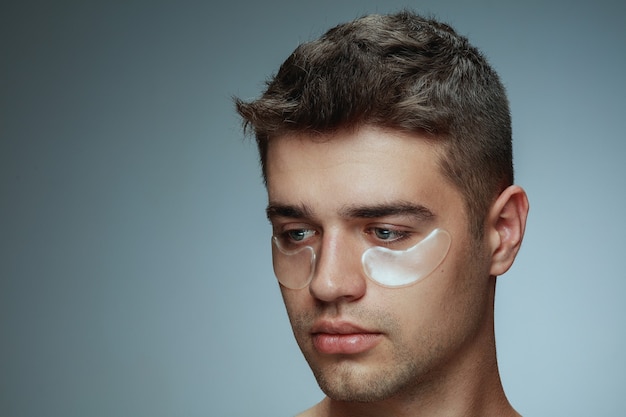 Het portret van het close-upprofiel van de jonge mens die op grijze studioachtergrond wordt geïsoleerd. Mannelijk gezicht met collageenvlekken onder de ogen. Concept van de gezondheid en schoonheid van mannen, cosmetologie, lichaams- en huidverzorging. Anti-veroudering.