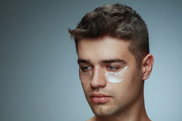 Het portret van het close-upprofiel van de jonge mens die op grijze studioachtergrond wordt geïsoleerd. Mannelijk gezicht met collageenvlekken onder de ogen. Concept van de gezondheid en schoonheid van mannen, cosmetologie, lichaams- en huidverzorging. Anti-veroudering.