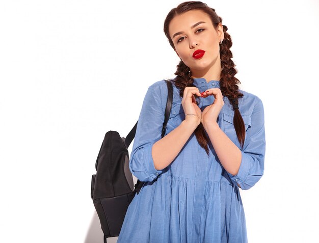 Het portret van jong gelukkig het glimlachen vrouwenmodel met heldere make-up en rode lippen met twee vlechten dient binnen geïsoleerde de zomer kleurrijke blauwe kleding en rugzak in. Hart teken tonen