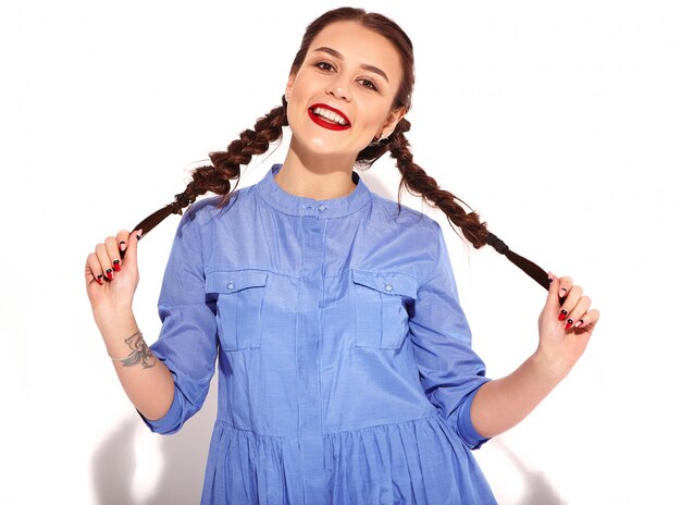 Het portret van jong gelukkig het glimlachen vrouwenmodel met heldere make-up en rode lippen met twee vlechten dient binnen geïsoleerde de zomer kleurrijke blauwe kleding in