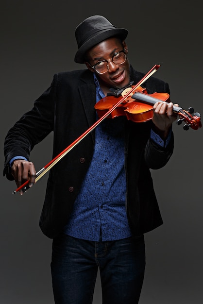 Het portret van knappe jonge zwarte lachende man in hoed viool spelen op donker