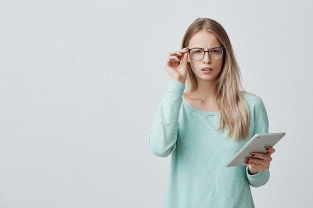 Het portret van ontevreden vermoeide jonge mooie onderneemster met blond haar in bril bevindt zich tegen grijze muur, werkt bij nieuw project op tablet, wil rust hebben. Negatieve emoties en gevoelens