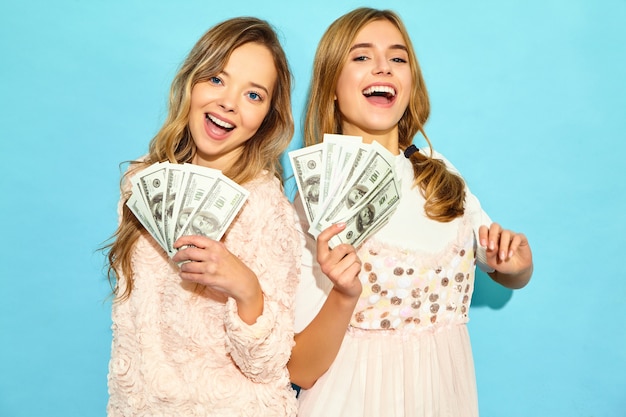 Het portret van twee gelukkige opgetogen blonde vrouwen die zich de zomerkleren verheugen verheugen zich en het houden van contant geldgeld dat over blauwe muur wordt geïsoleerd