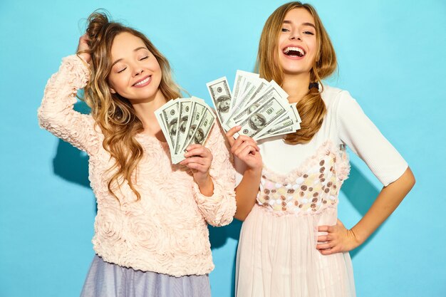 Het portret van twee gelukkige opgetogen blonde vrouwen die zich de zomerkleren verheugen verheugen zich en het houden van contant geldgeld dat over blauwe muur wordt geïsoleerd