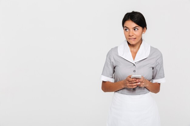 Het portret van vrij jonge donkerbruine vrouw in eenvormig bijt één lip terwijl het gebruiken van telefoon