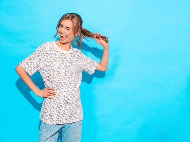 Het positieve vrouwelijke glimlachen. het grappige model stellen dichtbij blauwe muur in studio. het spelen met haar en knipoogt