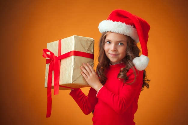 Het schattige vrolijke meisje met kerstmuts en cadeau op een oranje achtergrond