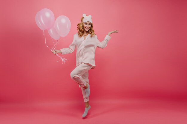 Het schot van gemiddelde lengte van feestvarken in het grijze sokken stellen. Portret van lachende jonge dame in zijden pyjama's springen met roze ballonnen.