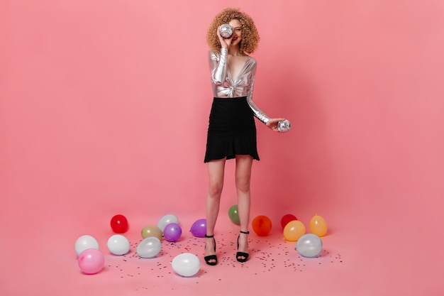 Het schot van gemiddelde lengte van krullend meisje in zilveren blouse en rok die discoballen op roze ruimte met ballons houden.