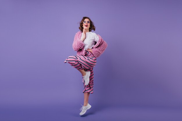 Het schot van gemiddelde lengte van lachend prachtig meisje in het witte sneakers springen. Foto van tevreden donkerbruine vrouw in roze kledij die zich op één been bevindt.