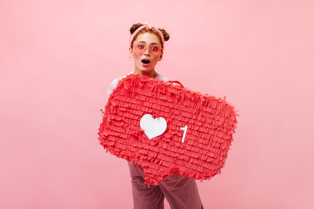 Het schot van gemiddelde lengte van mooie vrouw in overall en glazen die teken houden zoals op roze achtergrond.