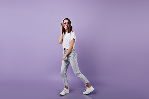 Het schot van gemiddelde lengte van slank meisje in jeans het luisteren muziek in hoofdtelefoons. Portret van vrouwelijk model in witte sneakers dansen.