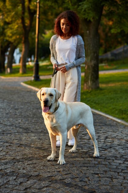 Het schot van gemiddelde lengte van vrij gezonde jonge dame die in de ochtend in park met hond loopt