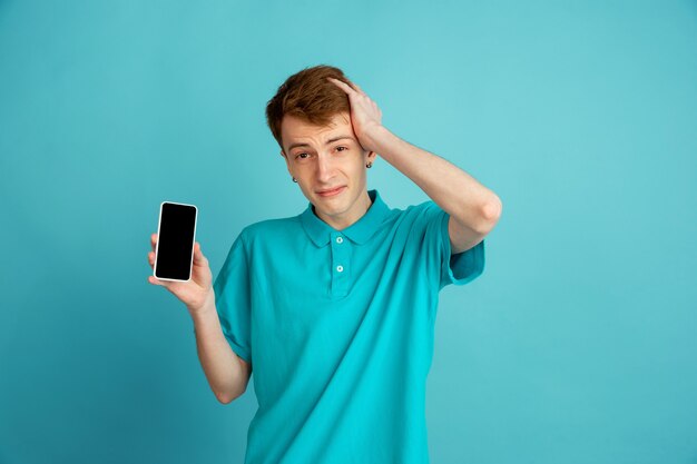 Het tonen van een leeg telefoonscherm. Het moderne portret van de blanke jongeman geïsoleerd op een blauwe muur, zwart-wit. Mooi mannelijk model. Concept van menselijke emoties, gezichtsuitdrukking, verkoop, advertentie, trendy.