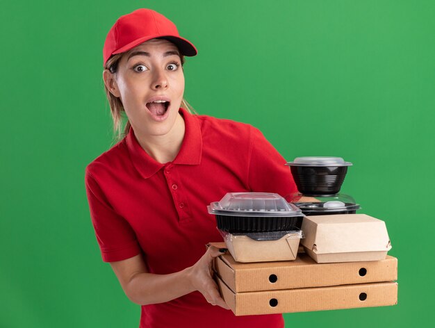 Het verbaasde jonge mooie bezorgmeisje in uniform houdt papieren voedselpakketten en containers op pizzadozen op groen