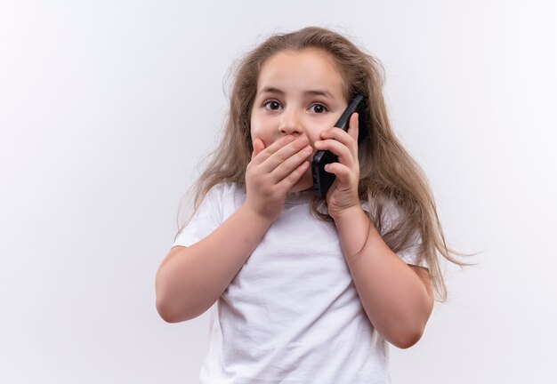 Het verbaasde kleine schoolmeisje die witte t-shirt dragen spreekt over telefoon behandelde mond op geïsoleerde witte achtergrond