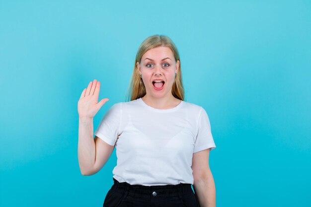 Het verraste blondemeisje toont hallo gebaar met hand op blauwe achtergrond