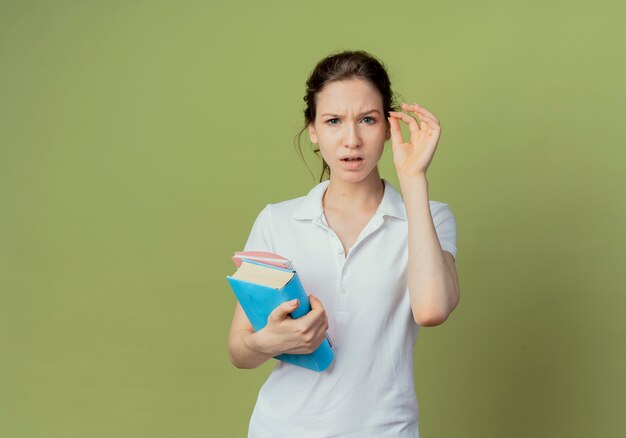 Het verwarde jonge mooie vrouwelijke boek en het notitieblok van de studentenholding dienen hand in lucht te houden alsof houdend iets geïsoleerd op olijfgroene achtergrond met exemplaarruimte