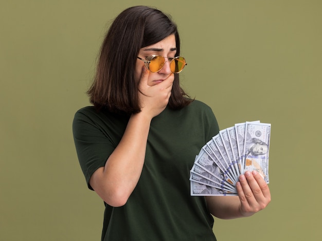 Het verwarde jonge vrij kaukasische meisje in zonnebril legt hand op kinholding en bekijkt geld op olijfgroen