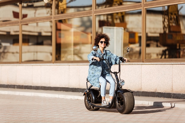 Het volledige lengtebeeld van glimlachende krullende vrouw in zonnebril berijdt in openlucht op moderne motor