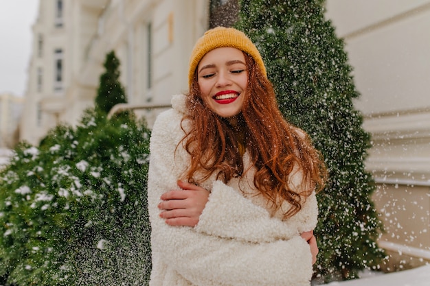 Het vrolijke gemberdame stellen onder sneeuw. glimlachende lieve vrouw in gebreide muts die zich dichtbij sparren bevindt.