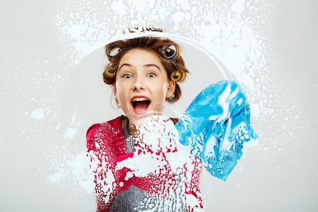 Het vrolijke jonge meisje wast vensters met blauwe handdoek