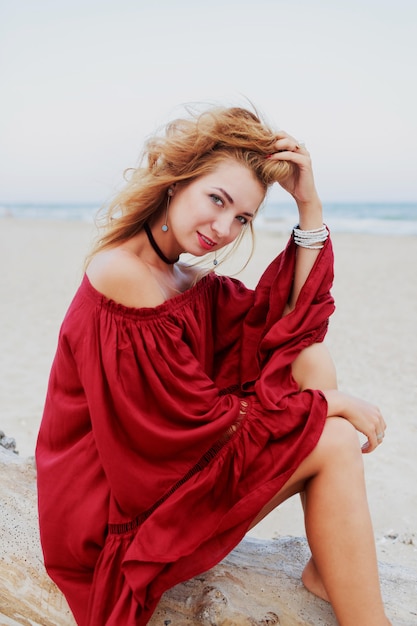 Het vrolijke red-head meisje stellen op strand. Zittend op wit zand. Winderige haren. Stijlvolle outfit. Lifestyle portret. Reisstemming. Oceaan kust.