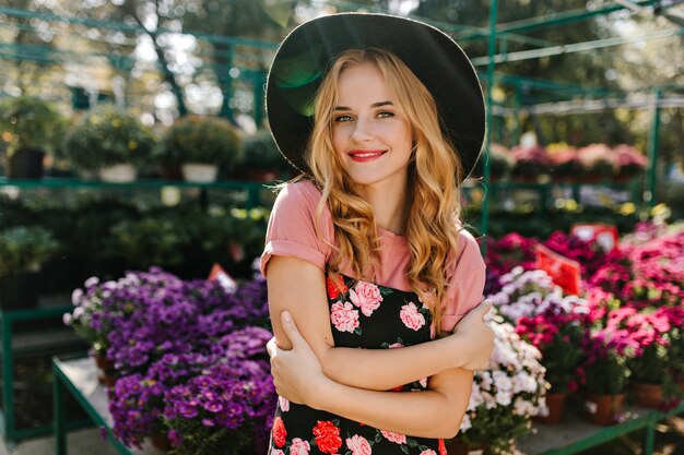 Het winnende blinde vrouw stellen op de oranjerie. Indoor portret van aangename blanke vrouw in zwarte hoed staande naast kleurrijke bloemen.