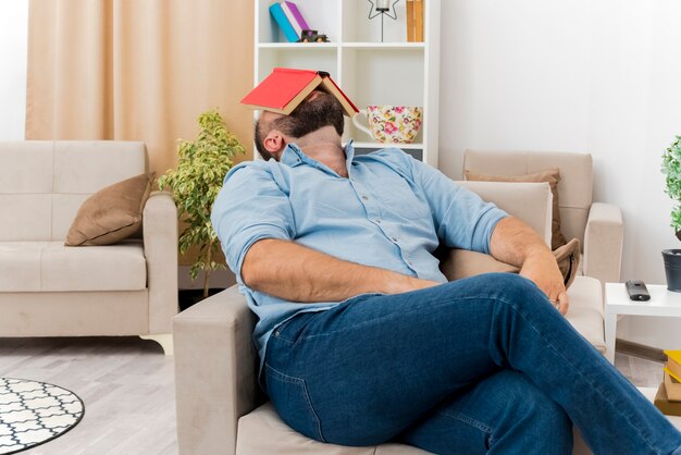 Het zetten van een volwassen Slavische man zit op een leunstoel met een boek op het gezicht in de woonkamer