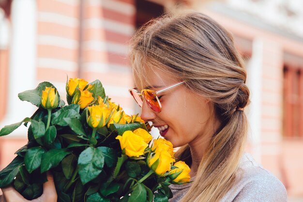 Het zijaanzicht van vrolijke schitterende vrouw in zonnebril, snuift gele rozen, buiten.