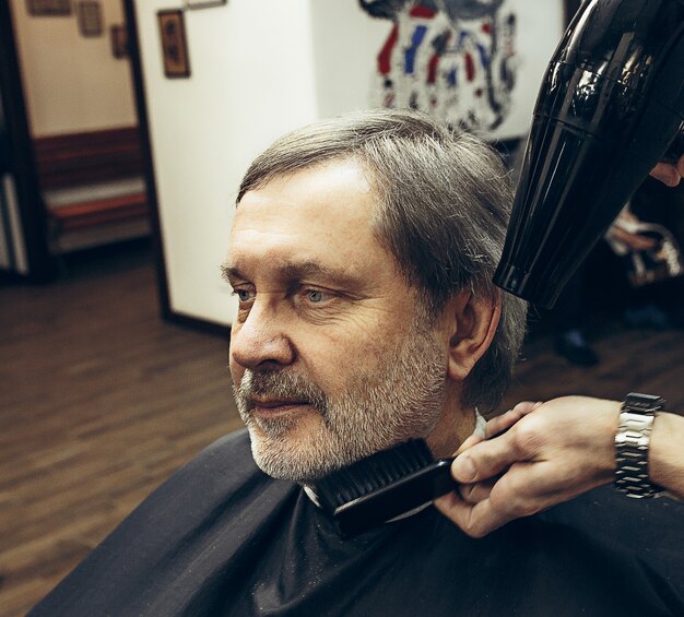 Het zijaanzichtportret van de close-up van de knappe hogere gebaarde Kaukasische mens die baard het verzorgen in moderne herenkapper krijgen.