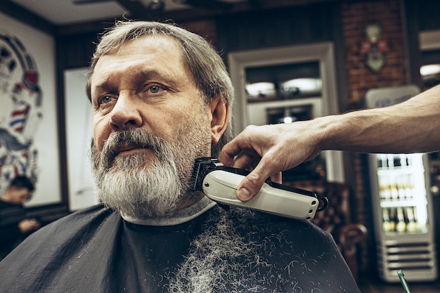 Het zijaanzichtportret van de close-up van de knappe hogere gebaarde Kaukasische mens die baard het verzorgen in moderne herenkapper krijgt.