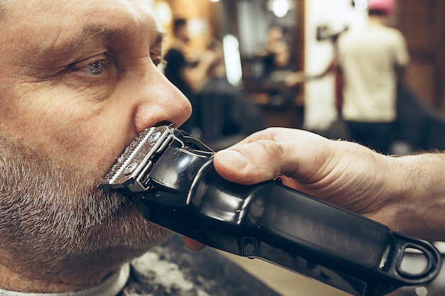 Het zijaanzichtportret van de close-up van de knappe hogere gebaarde Kaukasische mens die baard het verzorgen in moderne herenkapper krijgt.