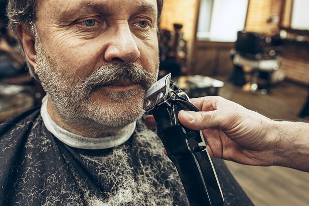 Het zijaanzichtportret van de close-up van de knappe hogere gebaarde Kaukasische mens die baard het verzorgen in moderne herenkapper krijgt.