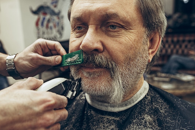 Het zijaanzichtportret van de close-up van de knappe hogere gebaarde Kaukasische mens die baard het verzorgen in moderne herenkapper krijgt.