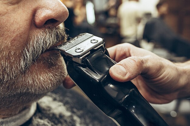 Het zijaanzichtportret van de close-up van de knappe hogere gebaarde Kaukasische mens die baard het verzorgen in moderne herenkapper krijgt.