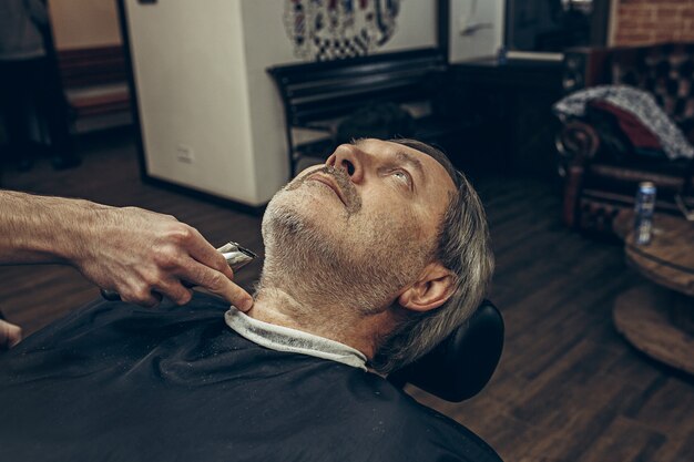 Het zijaanzichtportret van het close-up zijprofiel van knappe hogere gebaarde Kaukasische mens die baard het verzorgen in moderne herenkapper krijgen.