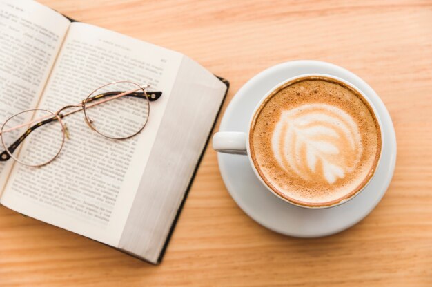 Hete koffiekop met cappuccino latte kunst en oogglazen over een open boek op tafel