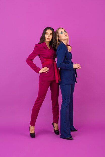 High fashion stijl twee lachende aantrekkelijke vrouwen op violette muur in stijlvolle kleurrijke avondpakken van paarse en blauwe kleur, vrienden samen plezier, modetrend