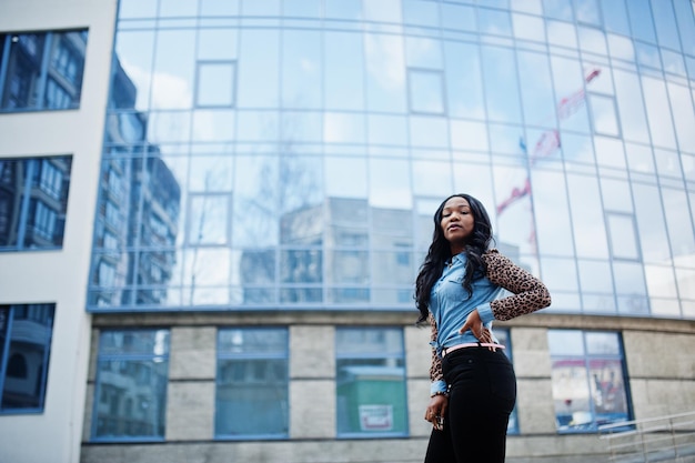 Hipster afrikaans amerikaans meisje met een spijkerbroek met luipaardmouwen die op straat poseren tegen een modern kantoorgebouw met blauwe ramen