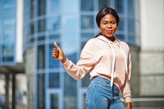 Hipster afrikaans amerikaans meisje met roze hoodie-jeans die zich voordeed op straat tegen kantoorgebouw met blauwe ramen
