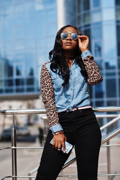 Hipster afro-amerikaans meisje met een zonnebril, jeans-shirt met luipaardmouwen, houdt een mobiele telefoon op straat tegen een modern kantoorgebouw