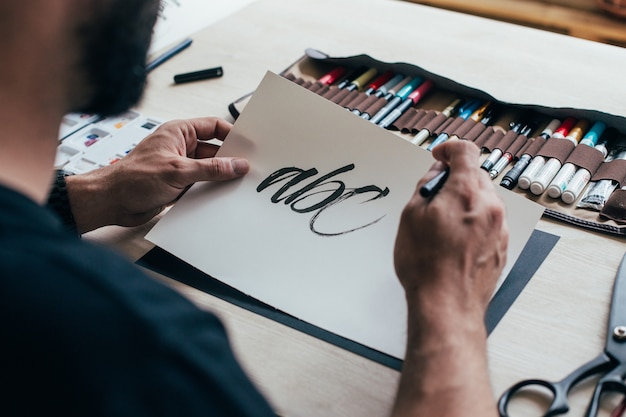 Gratis foto hipster jonge illustratorkunstenaar in zwarte, eenvoudige t-shirt creëert authentieke en unieke handbeletteringstekeningen in zijn lichte industriële studio