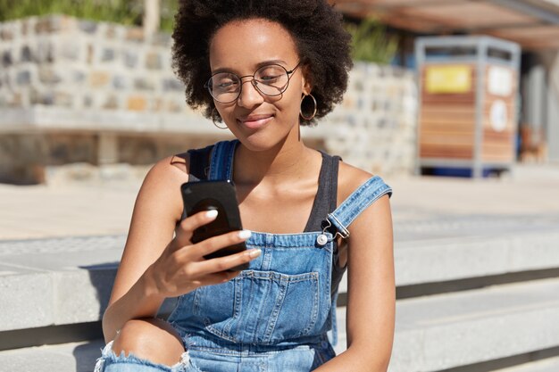 Hipster met donkere huid, knapperig haar, ontvangt sms op mobiele telefoon, draagt denim tuinbroek, optische bril, ronde oorbellen, bekijkt video op internet, ontspant buiten, maakt online winkelen