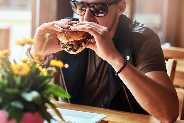 Hipster met een stijlvol kapsel en baard zit aan een tafel, besloot te dineren in een café langs de weg, een hamburger etend.