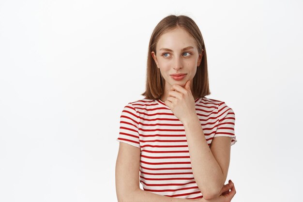 Hmm interessant. Jonge mooie vrouw denken, wegkijken met lachend nadenkend gezicht en kin aanraken, nadenken over besluit, staande in t-shirt tegen witte achtergrond